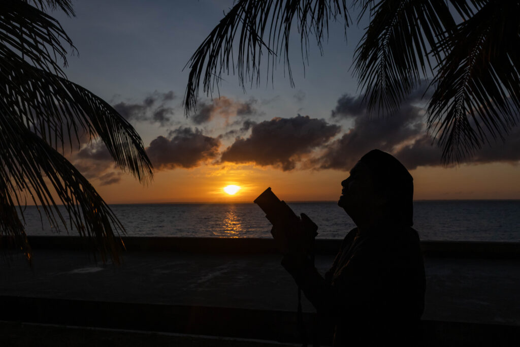 Fotógrafa em silhueta segurando a câmera diante do nascer do sol na orla de Macapá, com céu alaranjado e nuvens, emoldurada por folhas de palmeiras